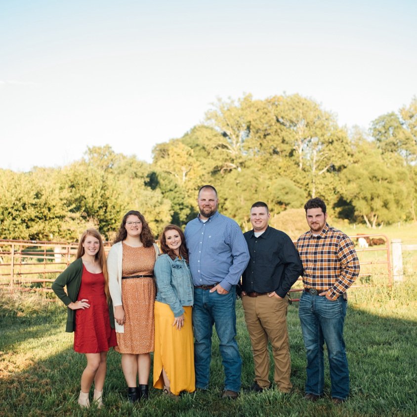 Group of six people standing together in a grassy field, smiling and dressed in casual attire. The background features trees and a fence, suggesting a rural or farm setting. Ideal for family or group portrait themes.